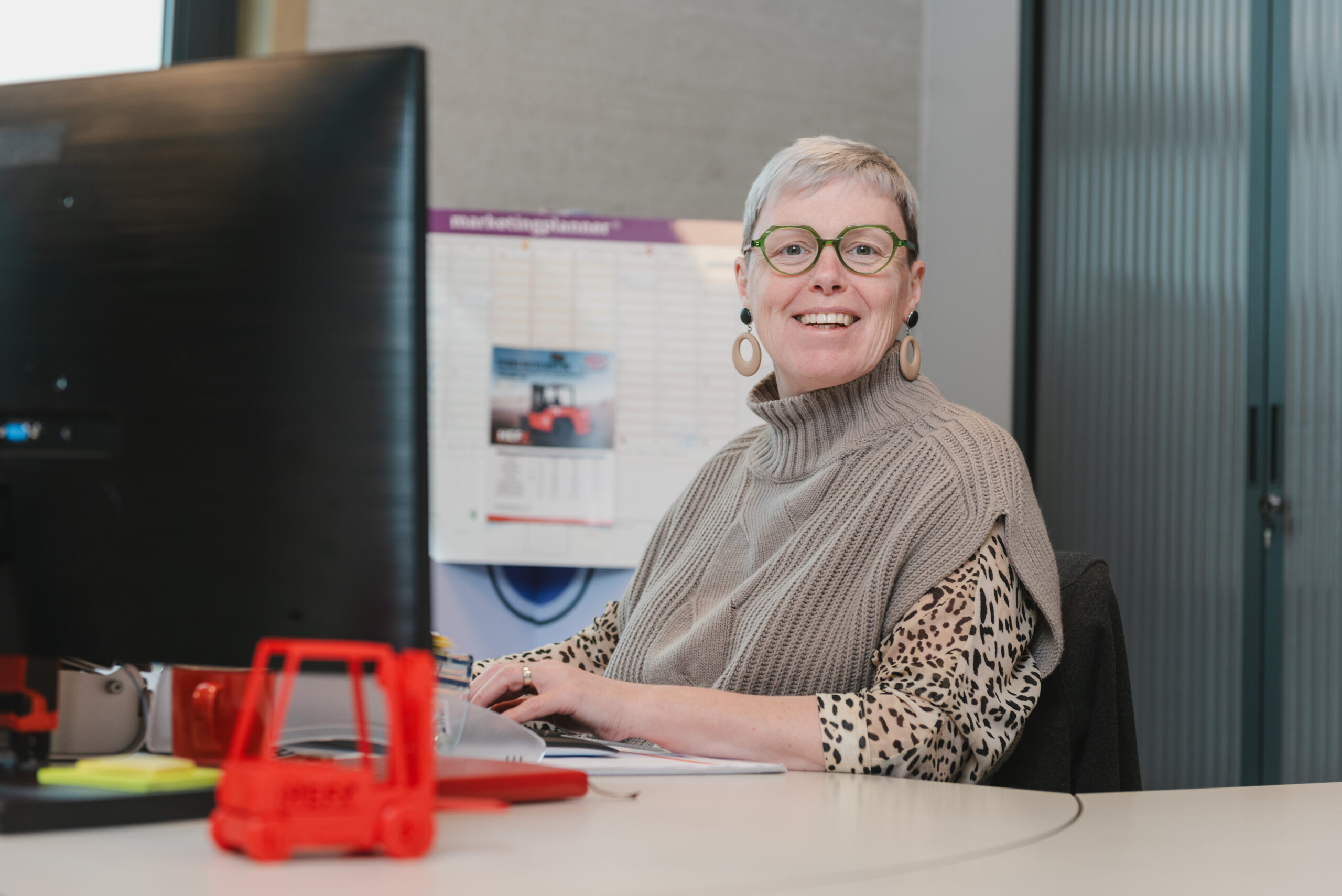 Sofie van HefX zit glimlachend achter een bureau met een groot zwart computerscherm. Ze draagt een mouwloos vest met een col over een blouse met dierenprint. Een rode speelgoedvorkheftruck staat op haar bureau.