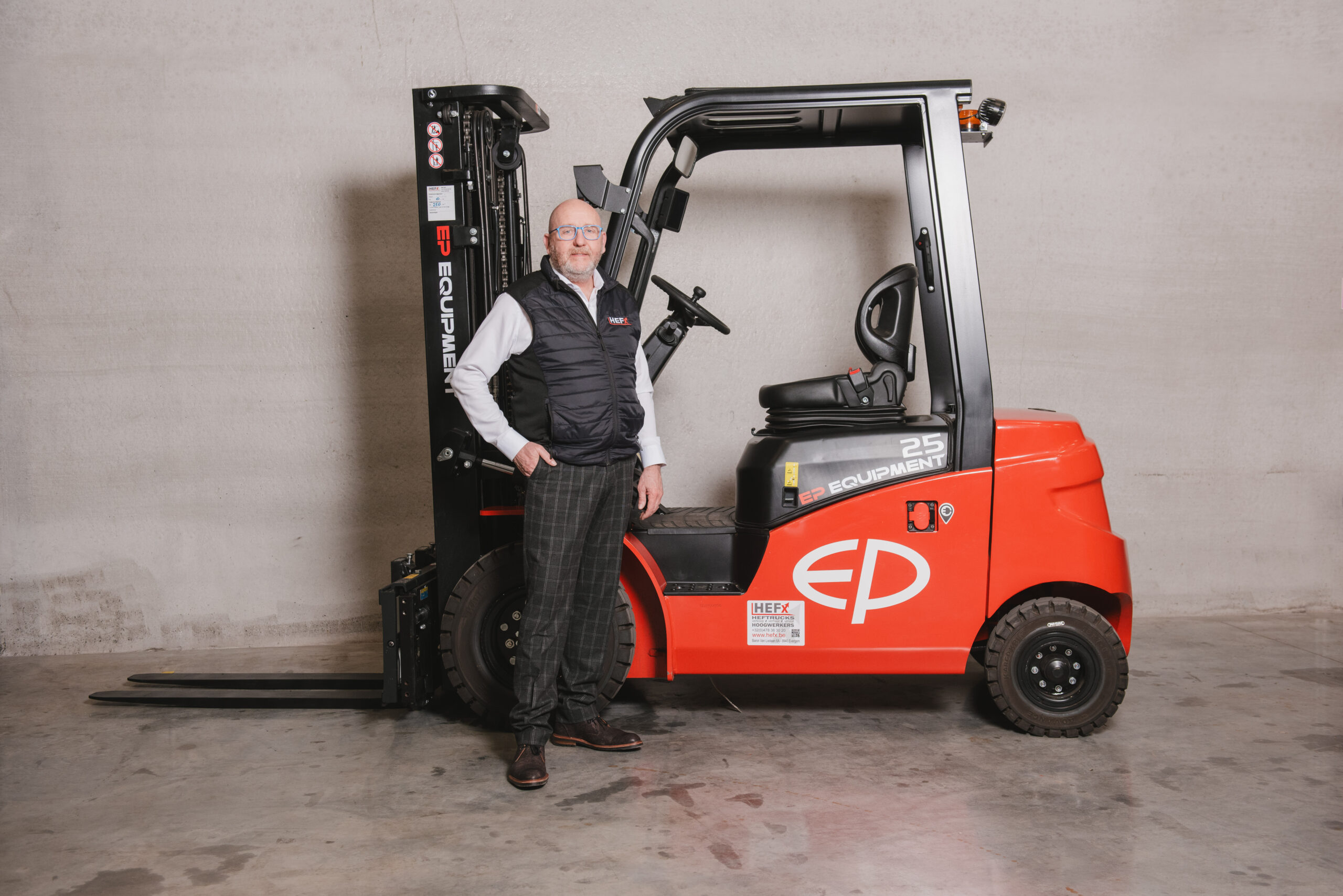 Man poseert voor een EP Equipment CPD25 elektrische heftruck in een industriële ruimte met betonnen vloer en neutrale achtergrond, voorzien van HEFX-label op het voertuig.