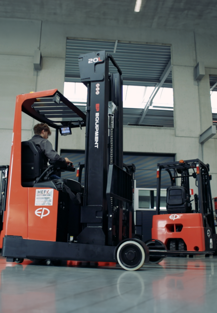 Chauffeur bestuurt een elektrische reachtruck van EP Equipment in een magazijnruimte, met focus op de hefmast en bedieningselementen van het voertuig.